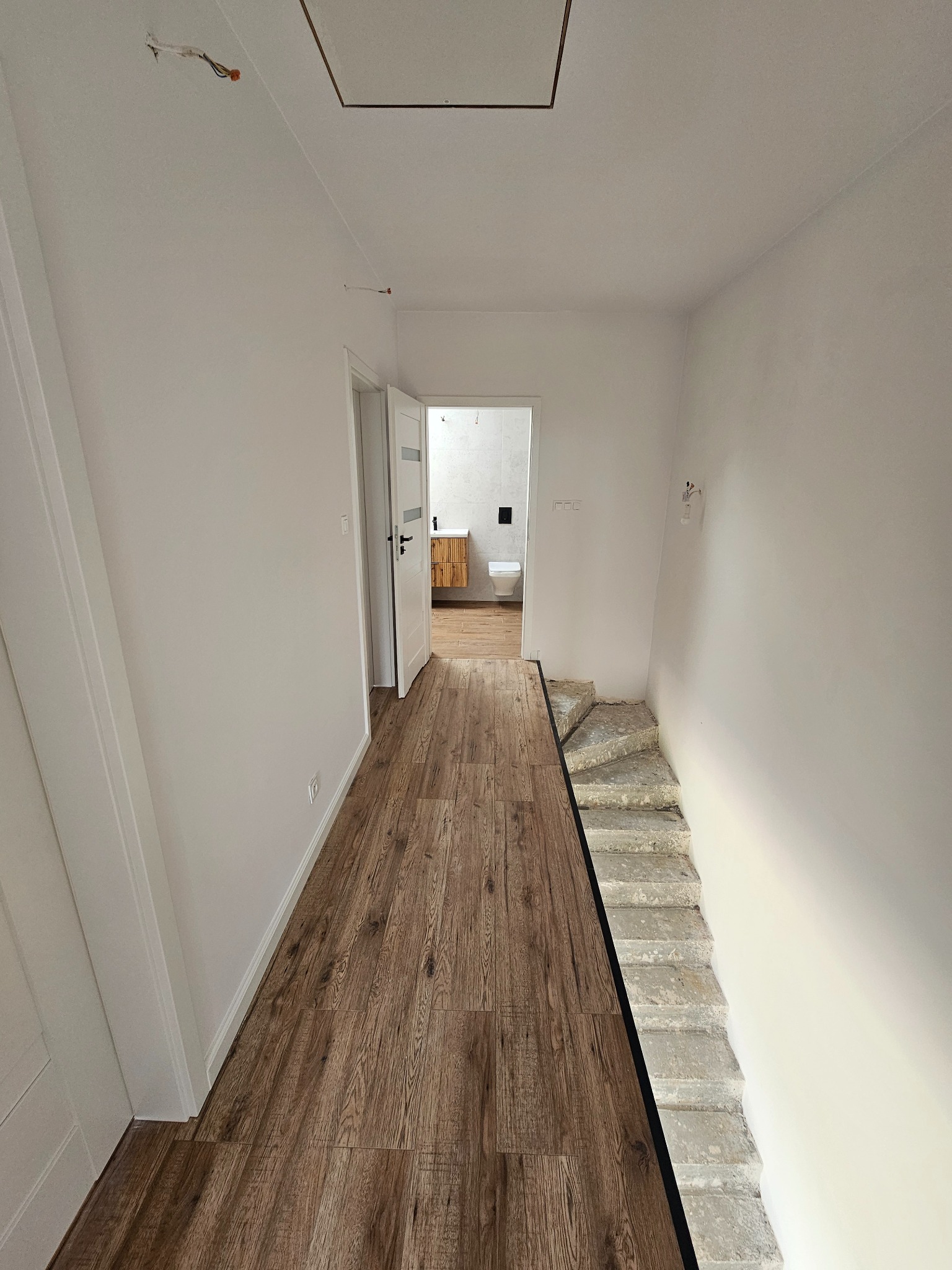 Korytarz z drewnopodobną podłogą, prowadzący do otwartych drzwi łazienki z widoczną toaletą i szafką z drewnianymi frontami; surowe betonowe schody obok podłogi, białe ściany z wystającymi przewodami.
