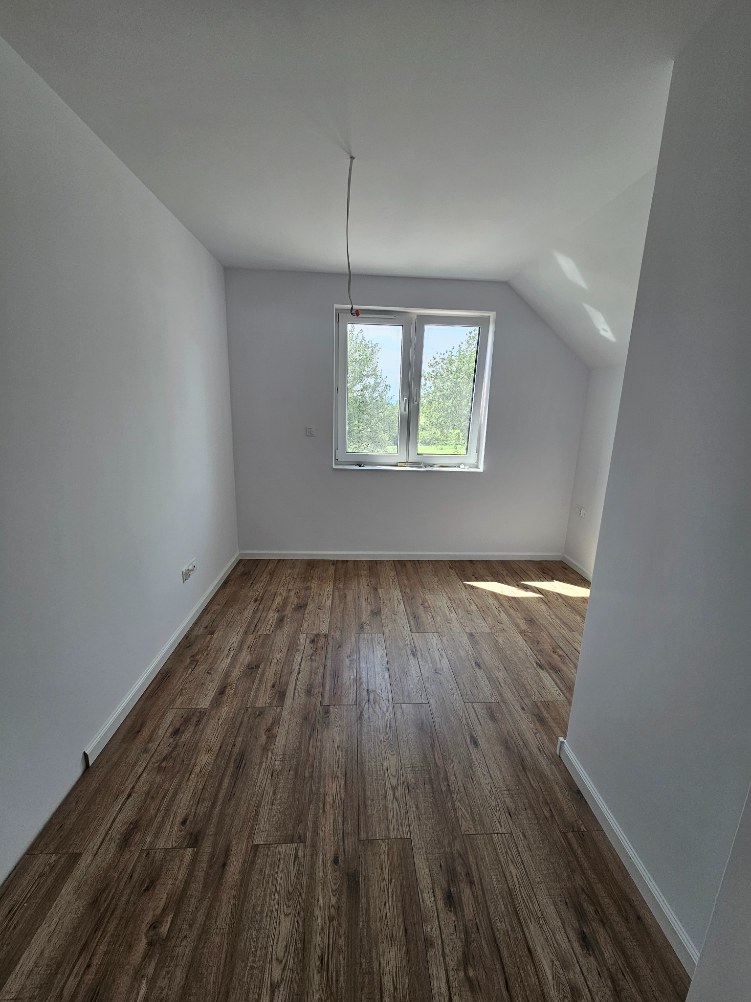 Pusty pokój na poddaszu z drewnianą podłogą, białymi ścianami, oknem z widokiem na zieleń i wystającym przewodem z sufitu.