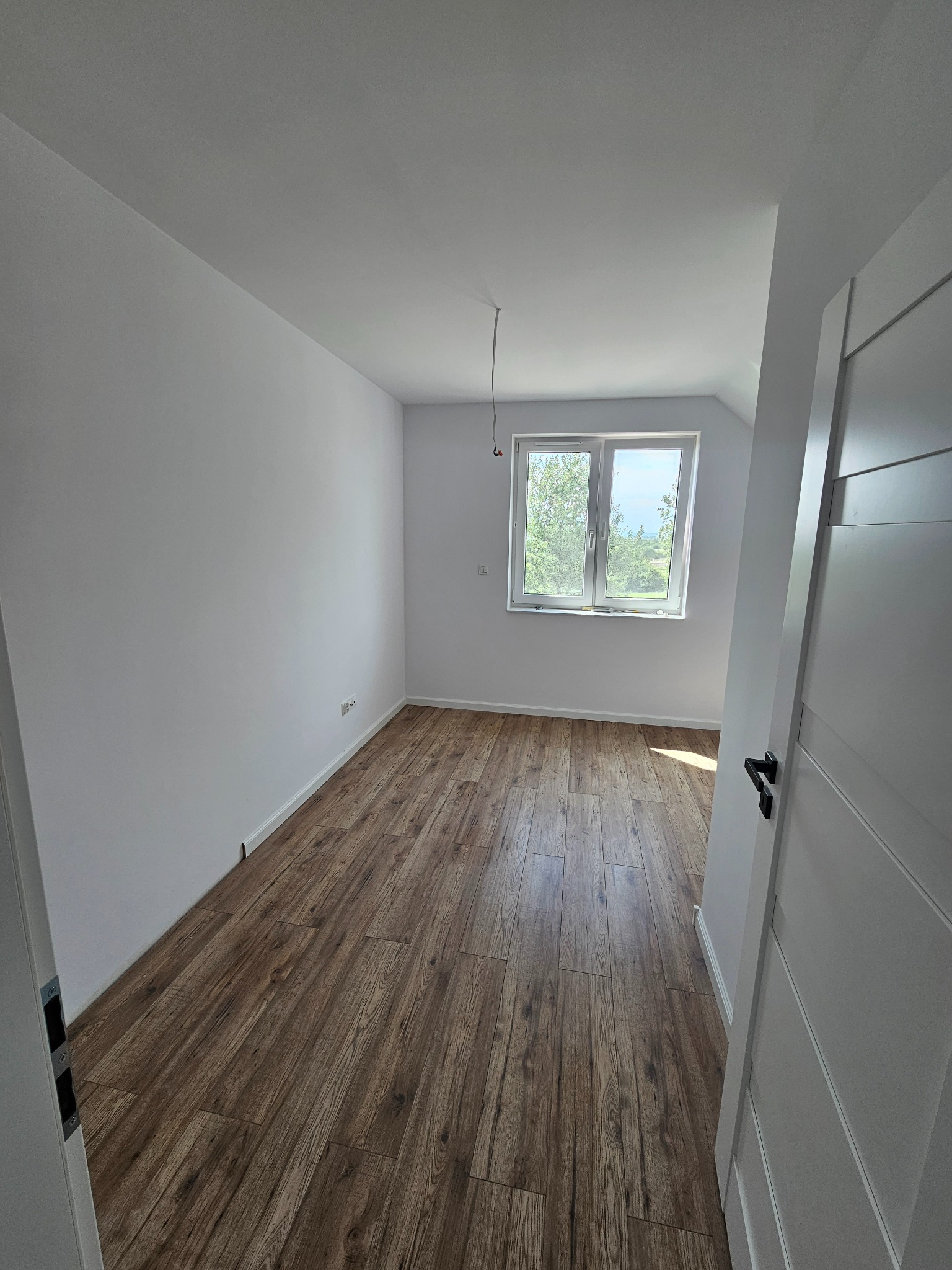 Pomalowane na biało wnętrze pokoju z drewnopodobną podłogą, widok na okno z zielenią na zewnątrz, białe ściany i częściowo widoczne białe drzwi z czarną klamką.