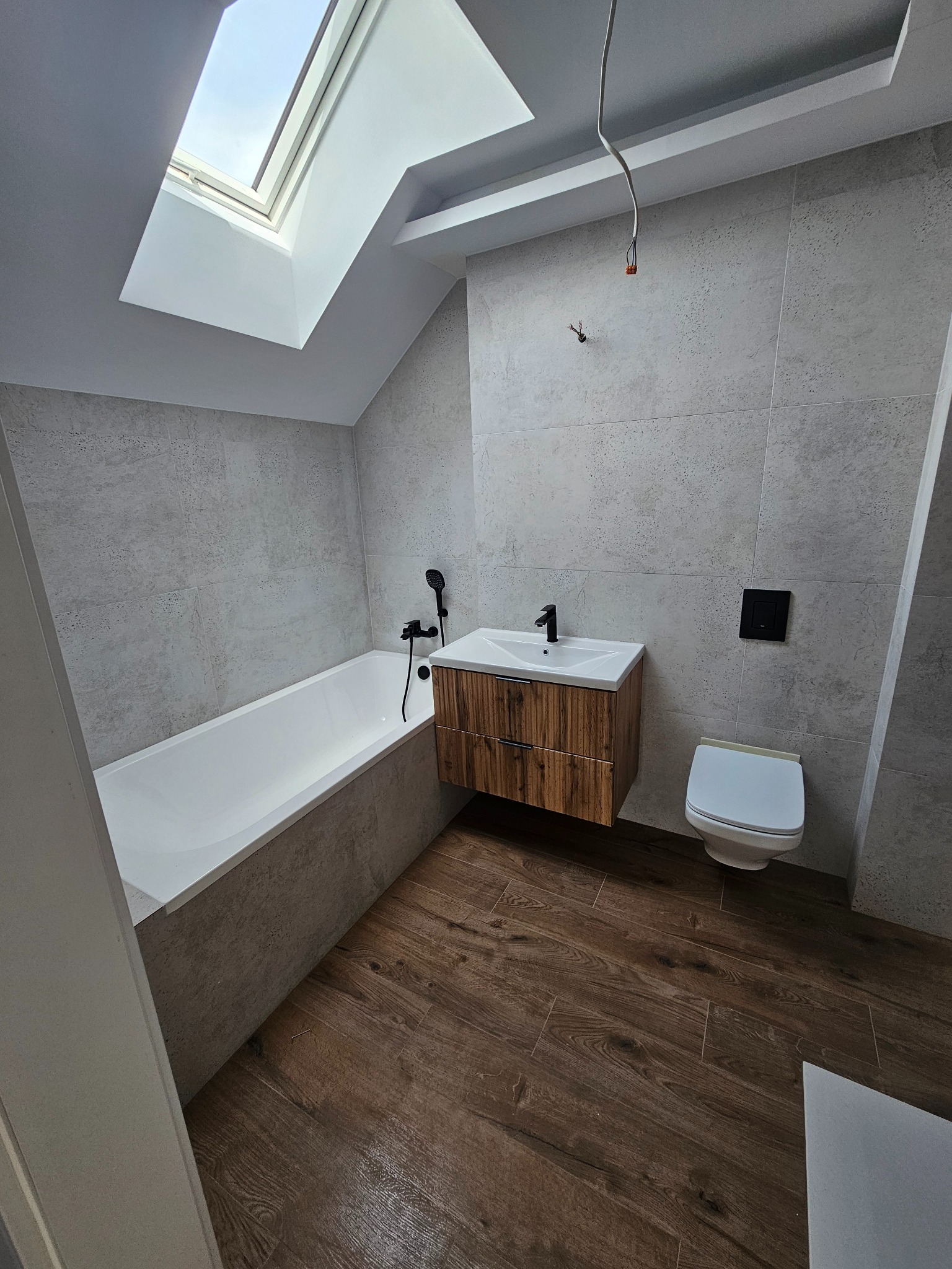 Nowoczesna łazienka na poddaszu z oknem dachowym, wykończona płytkami imitującymi beton i drewnem, z wiszącą szafką umywalkową i czarną armaturą, widoczne niedokończone podłączenie elektryczne.