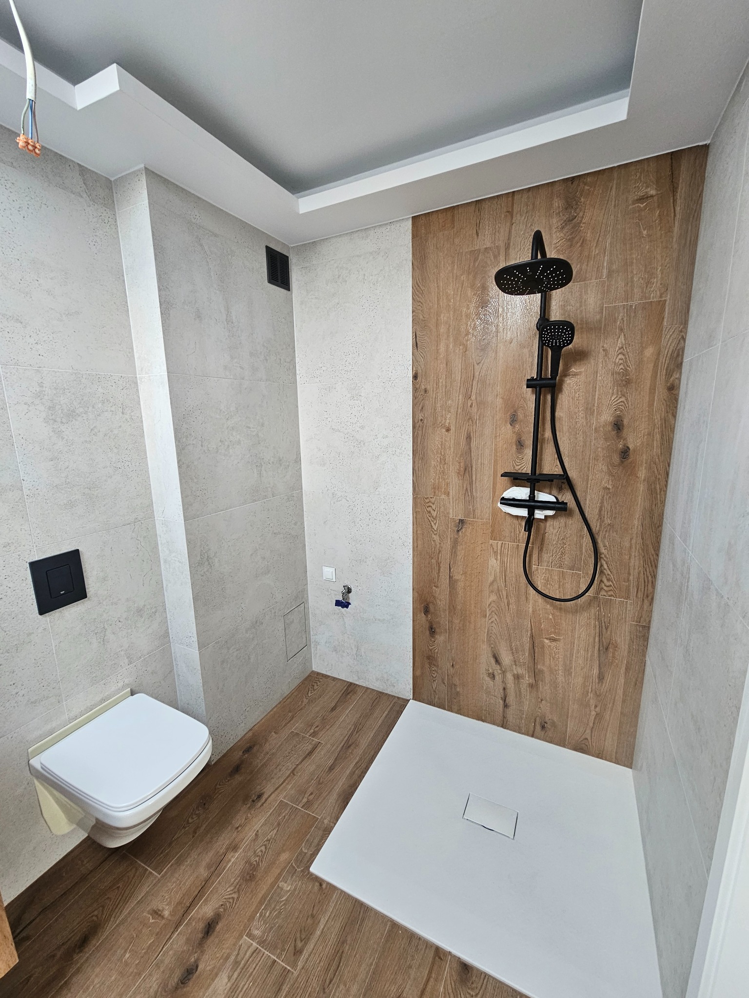 Nowoczesna łazienka po remoncie: kabina prysznicowa typu walk-in z czarną armaturą na tle drewnopodobnych płytek, podwieszana toaleta i płytki imitujące drewno na podłodze.