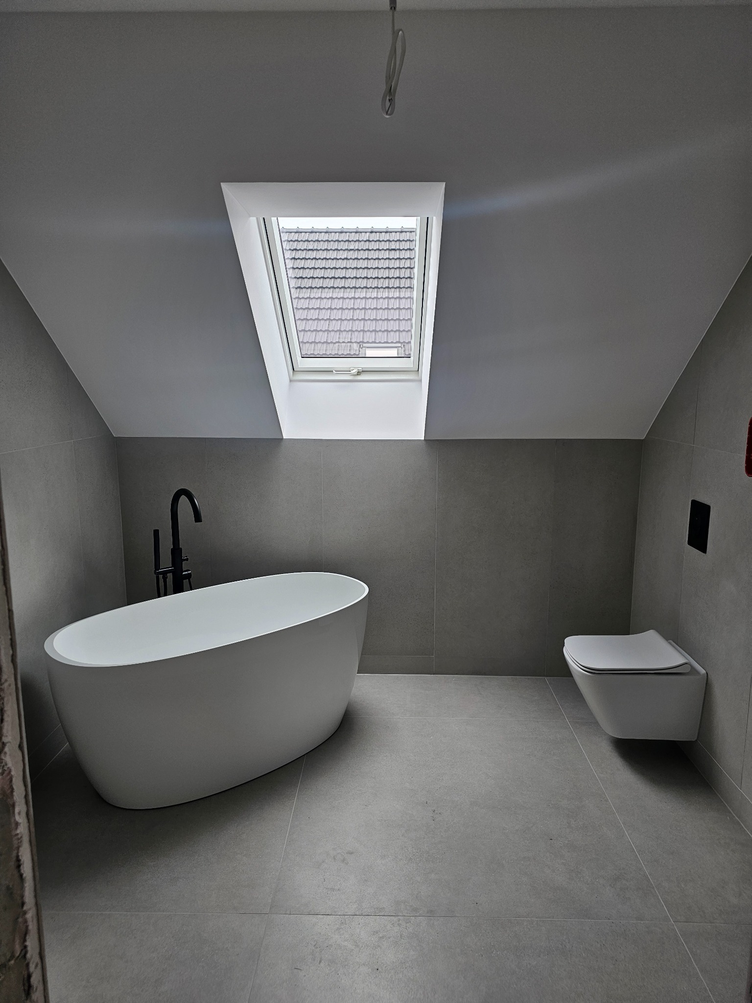 Nowoczesna łazienka na poddaszu z oknem dachowym, szare płytki ceramiczne, wolnostojąca wanna z czarną armaturą i podwieszana toaleta.