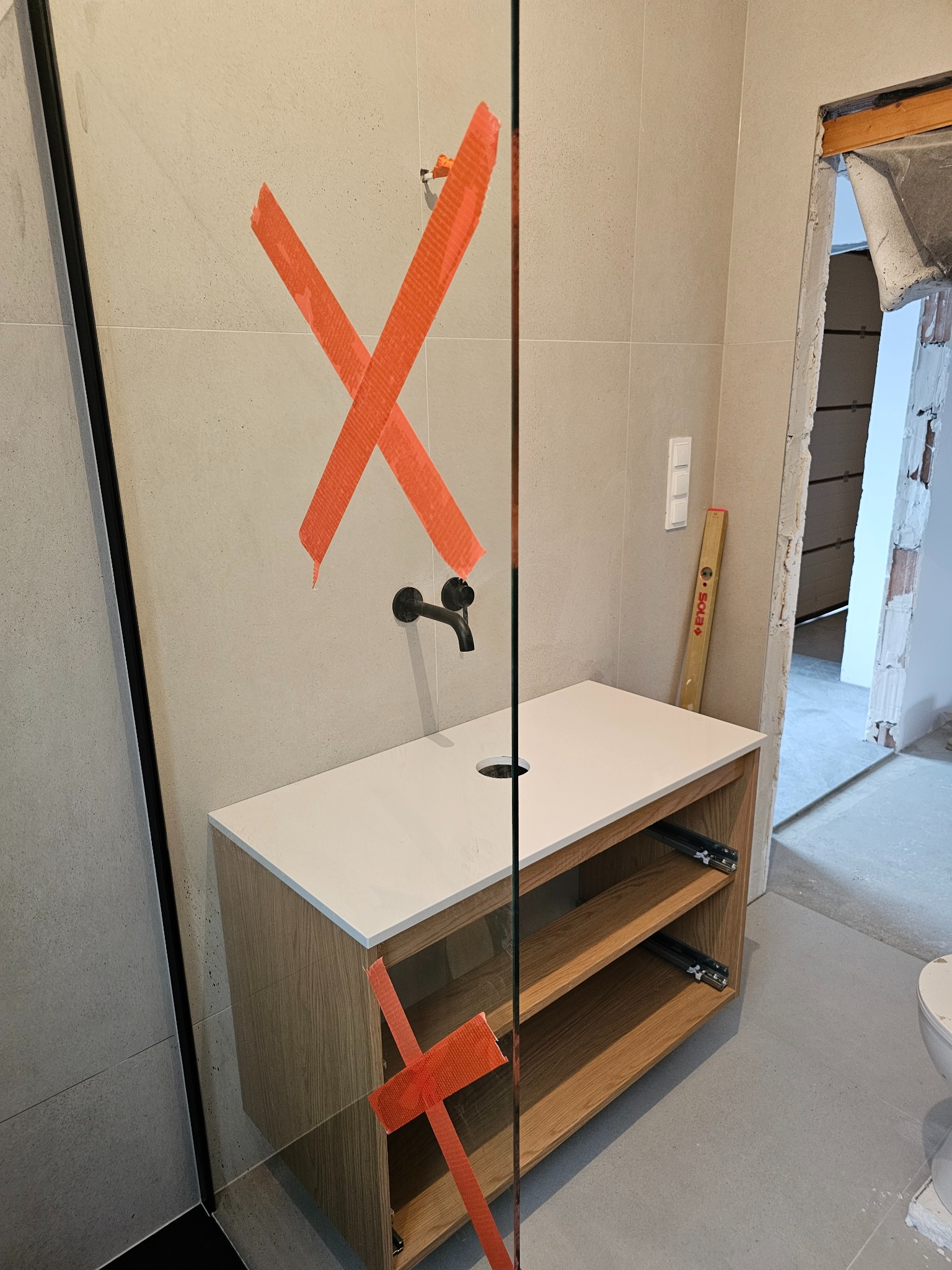 Niedokończona łazienka z umywalką nablatową, czarną armaturą i płytkami ściennymi w beżowym odcieniu, zabezpieczona pomarańczową taśmą malarską. W tle widoczny fragment nieotynkowanej ściany...