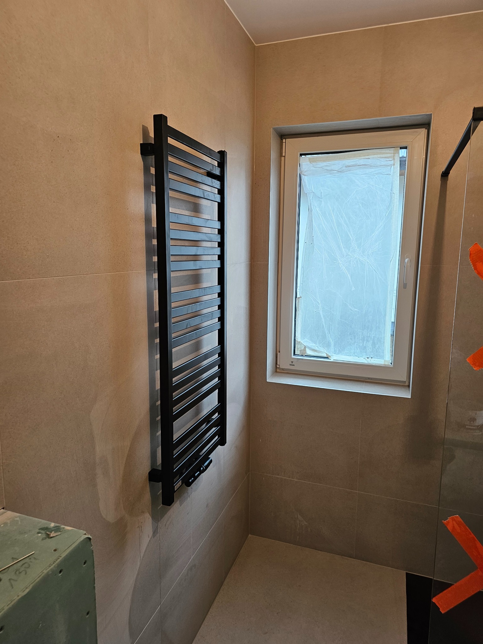 Nowoczesna łazienka w trakcie wykańczania: czarny grzejnik drabinkowy na ścianie z płytek w odcieniach beżu, okno zabezpieczone folią malarską, fragment kabiny prysznicowej z pomarańczową taśmą...