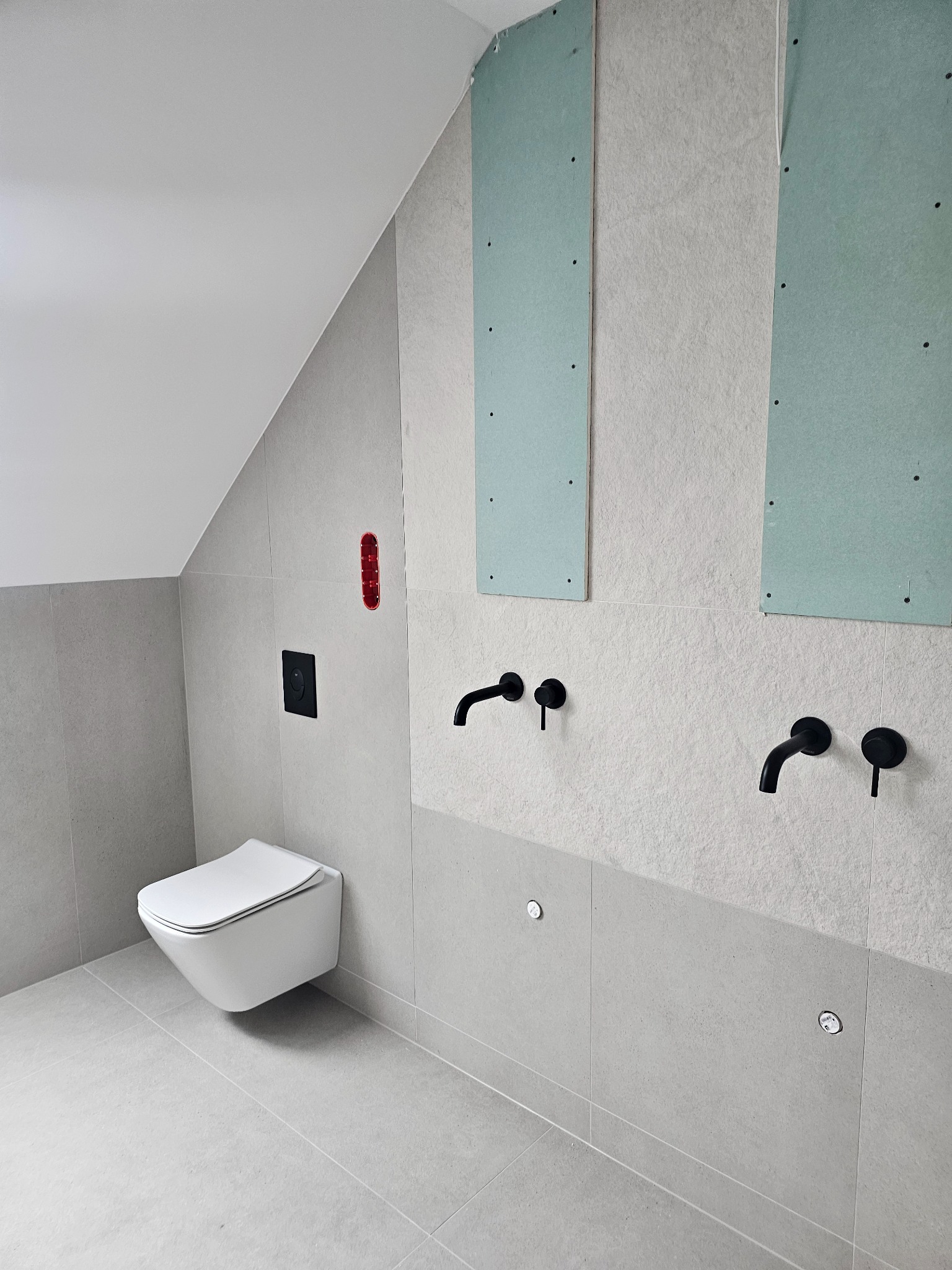 Wnętrze łazienki w Bolesławiu w trakcie remontu, z zamontowaną podtynkową toaletą, widoczne fragmenty płyt kartonowo-gipsowych na ścianie oraz czarne baterie umywalkowe.