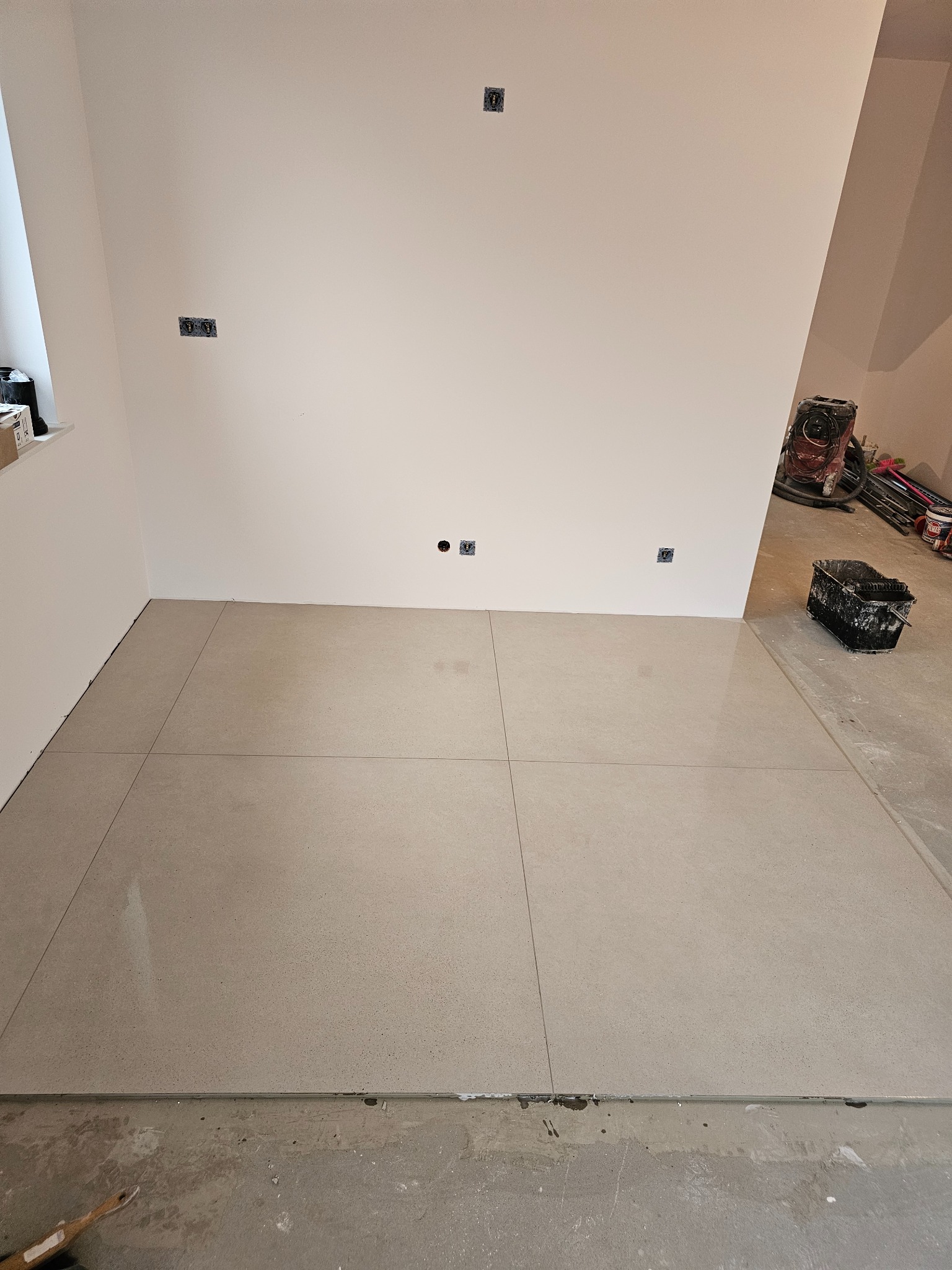 Ułożone jasne płytki podłogowe z fugami, widoczne gniazdka elektryczne na ścianie, narzędzia malarskie i szlifierka w tle, podłoga przed płytkami do wykończenia.