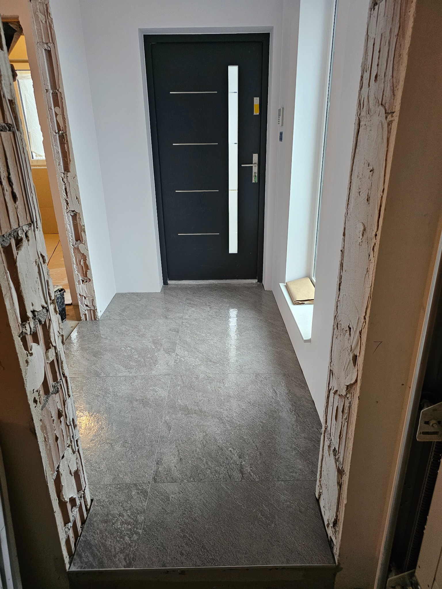Szare płytki podłogowe w przedpokoju, widoczne fragmenty ścian w trakcie prac wykończeniowych, czarne drzwi wejściowe z poziomymi srebrnymi paskami, parapet z brązowym pakunkiem, surowe wykończenie...