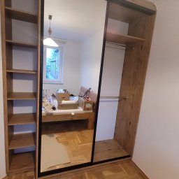 USŁUGI STOLARSKIE KOTECKI JACEK - Szafa wnękowa z przesuwnymi drzwiami lustrzanymi, z drewnianymi półkami po bokach, widoczna w trakcie montażu, z kartonami i narzędziami w tle, na parkiecie.