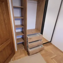 Wysoka szafa wnękowa z lustrem i otwartymi półkami, drewnopodobny dekor, trzy wysuwane szuflady z białymi wnętrzami, parkiet na podłodze częściowo zakryty kartonem ochronnym.