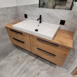 Nowoczesna łazienka z drewnianą szafką pod umywalkę z czterema szufladami i czarnymi uchwytami na tle marmurowych płytek.