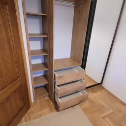 USŁUGI STOLARSKIE KOTECKI JACEK - Wysoka szafa wnękowa z przesuwnymi drzwiami lustrzanymi i otwartymi półkami, wykończona drewnopodobnym laminatem w ciepłym odcieniu dębu, z trzema wysuwanymi szufladami, widoczna na tle parkietu...