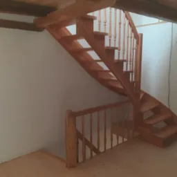 Drewniane, spiralne schody z balustradą, wykonane z jasnego drewna, umieszczone w jasnym pomieszczeniu z drewnianymi belkami stropowymi.