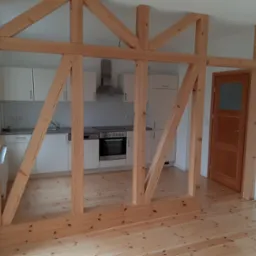 Drewniana konstrukcja szkieletowa oddzielająca kuchnię od salonu, widoczne elementy zabudowy kuchennej i drzwi wewnętrzne, jasna podłoga z desek.