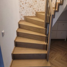 USŁUGI STOLARSKIE KOTECKI JACEK - Jasne drewniane schody ze szczeblami w ciemnym kolorze, widoczne na tle ściany z wzorem liści i parkietu ułożonego w jodełkę.