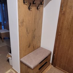 USŁUGI STOLARSKIE KOTECKI JACEK - Drewniany panel ścienny z wieszakami i półką w przedpokoju, połączony z siedziskiem z szufladami, widoczne niedokończone prace wykończeniowe podłogi.