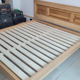 USŁUGI STOLARSKIE KOTECKI JACEK - Rama łóżka z jasnego drewna z prostym wezgłowiem, widoczne skrzynki narzędziowe i odkurzacz przemysłowy w tle, montaż w trakcie.