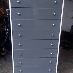 USŁUGI STOLARSKIE KOTECKI JACEK - Wysoka komoda z siedmioma szufladami w kolorze szarym, z białym obramowaniem i okrągłymi uchwytami, ustawiona na kółkach w garażu.