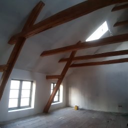 USŁUGI STOLARSKIE KOTECKI JACEK - Poddasze z drewnianą konstrukcją więźby dachowej, widoczne okna dachowe i ściany przygotowane do wykończenia, podłoga z desek. Na podłodze widoczne ślady po remoncie i narzędzia.