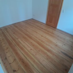 USŁUGI STOLARSKIE KOTECKI JACEK - Jasne, drewniane deski podłogowe ułożone w pokoju z białymi ścianami i drewnianymi drzwiami. Widoczne ślady użytkowania i naturalna struktura drewna.