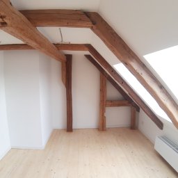 USŁUGI STOLARSKIE KOTECKI JACEK - Poddasze z widocznymi elementami konstrukcyjnymi dachu: drewnianymi belkami nośnymi, słupami i jasną drewnianą podłogą. Ściany białe, z oknem dachowym wpadającym w kadr. Widoczny grzejnik pod oknem.