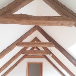 USŁUGI STOLARSKIE KOTECKI JACEK - Drewniana konstrukcja dachu z widocznymi belkami w jasnym, odnowionym wnętrzu poddasza, z surowym wykończeniem i żarówką na kablu.