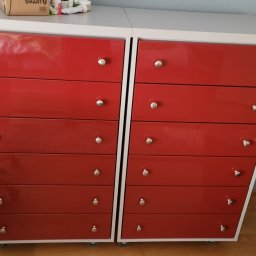 USŁUGI STOLARSKIE KOTECKI JACEK - Dwie czerwone komody z sześcioma szufladami każda, z białymi blatami i bokami, na kółkach, z okrągłymi, srebrnymi uchwytami, ustawione obok siebie na drewnianej podłodze.