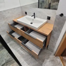 Nowoczesna szafka łazienkowa z drewnianym blatem i umywalką nablatową; otwarte szuflady z szarymi wnętrzami; marmurowe płytki na ścianach i podłodze; widoczny fragment kabiny prysznicowej...