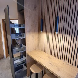 USŁUGI STOLARSKIE KOTECKI JACEK - Drewniany blat barowy z dwoma hokerami na czarnych nogach, w tle ściana z drewnianych paneli i czarne lampy wiszące, obok szafka cargo z wysuwanymi półkami.