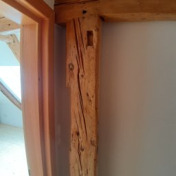 USŁUGI STOLARSKIE KOTECKI JACEK - Surowe drewno konstrukcyjne tworzące framugę drzwi, kontrastujące z gładką, pomalowaną ścianą w tle, widoczne ślady obróbki na belce.