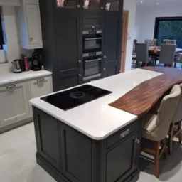 Nowoczesna kuchnia z wyspą, ciemnoszare szafki z przeszklonymi górnymi frontami, biały blat wyspy z wbudowaną płytą indukcyjną i drewnianym blatem barowym, widoczne krzesła barowe i stół jadalny...