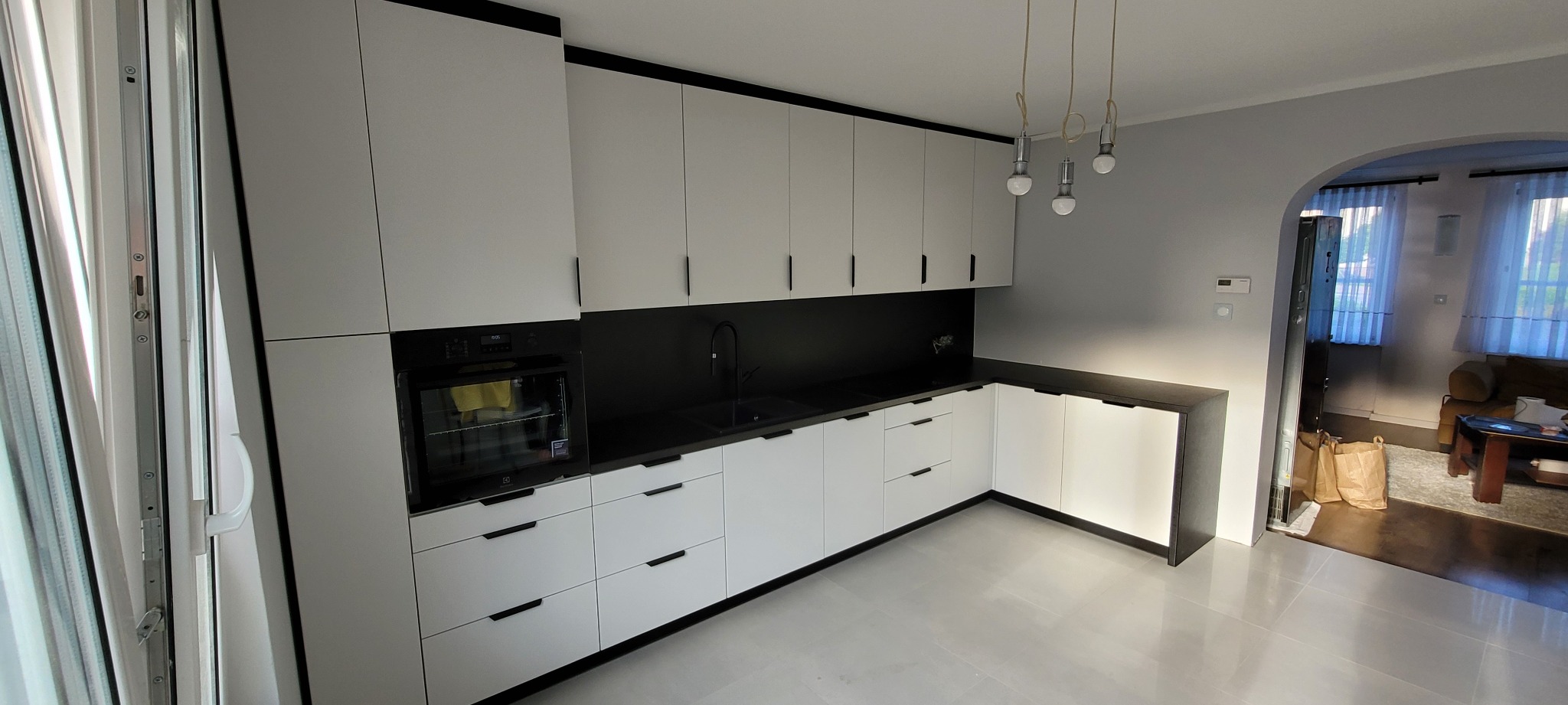 Nowoczesna kuchnia z białymi frontami, czarnym blatem i subtelnym oświetleniem punktowym, widok na salon przez przeszklone drzwi przesuwne.