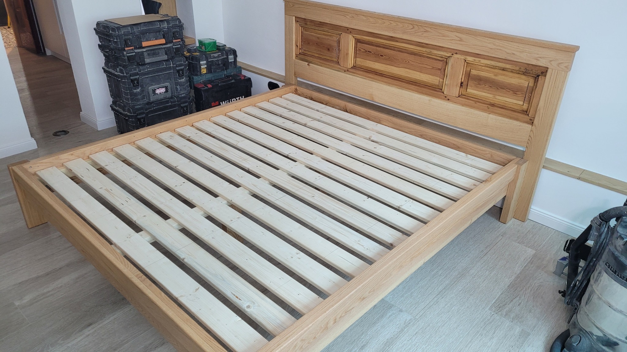 Rama łóżka z jasnego drewna z prostym wezgłowiem, widoczne skrzynki narzędziowe i odkurzacz przemysłowy w tle, montaż w trakcie.