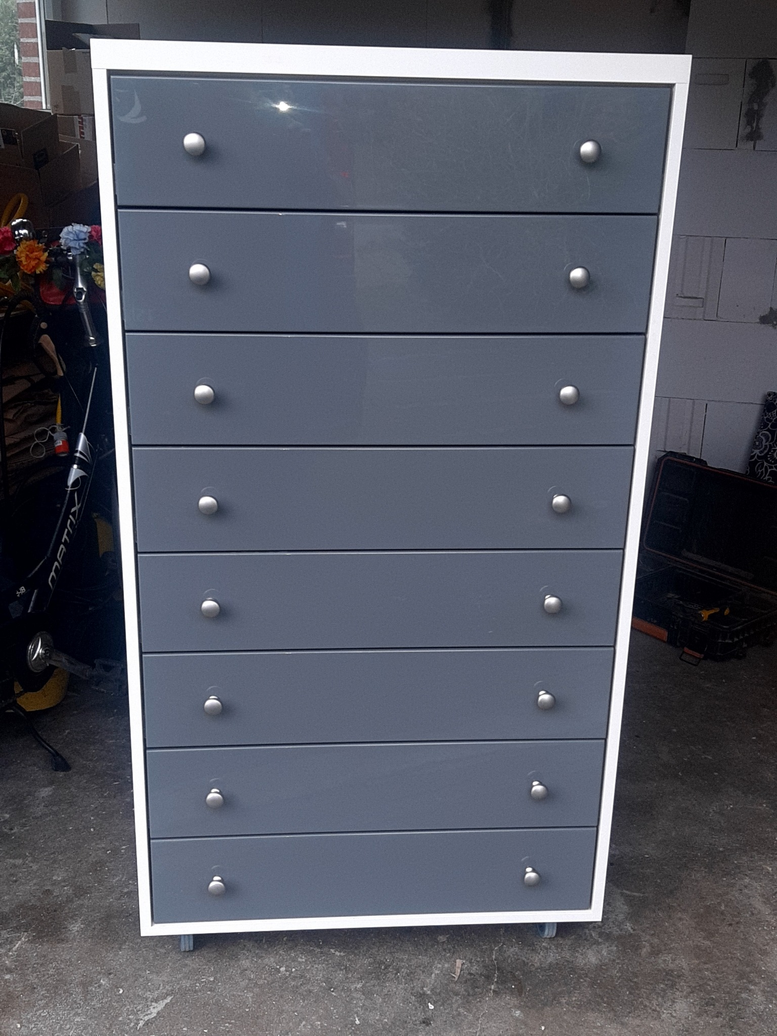 Wysoka komoda z siedmioma szufladami w kolorze szarym, z białym obramowaniem i okrągłymi uchwytami, ustawiona na kółkach w garażu.