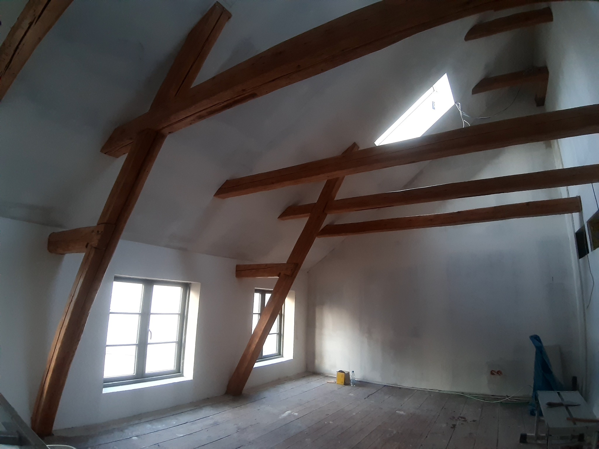 Poddasze z drewnianą konstrukcją więźby dachowej, widoczne okna dachowe i ściany przygotowane do wykończenia, podłoga z desek. Na podłodze widoczne ślady po remoncie i narzędzia.