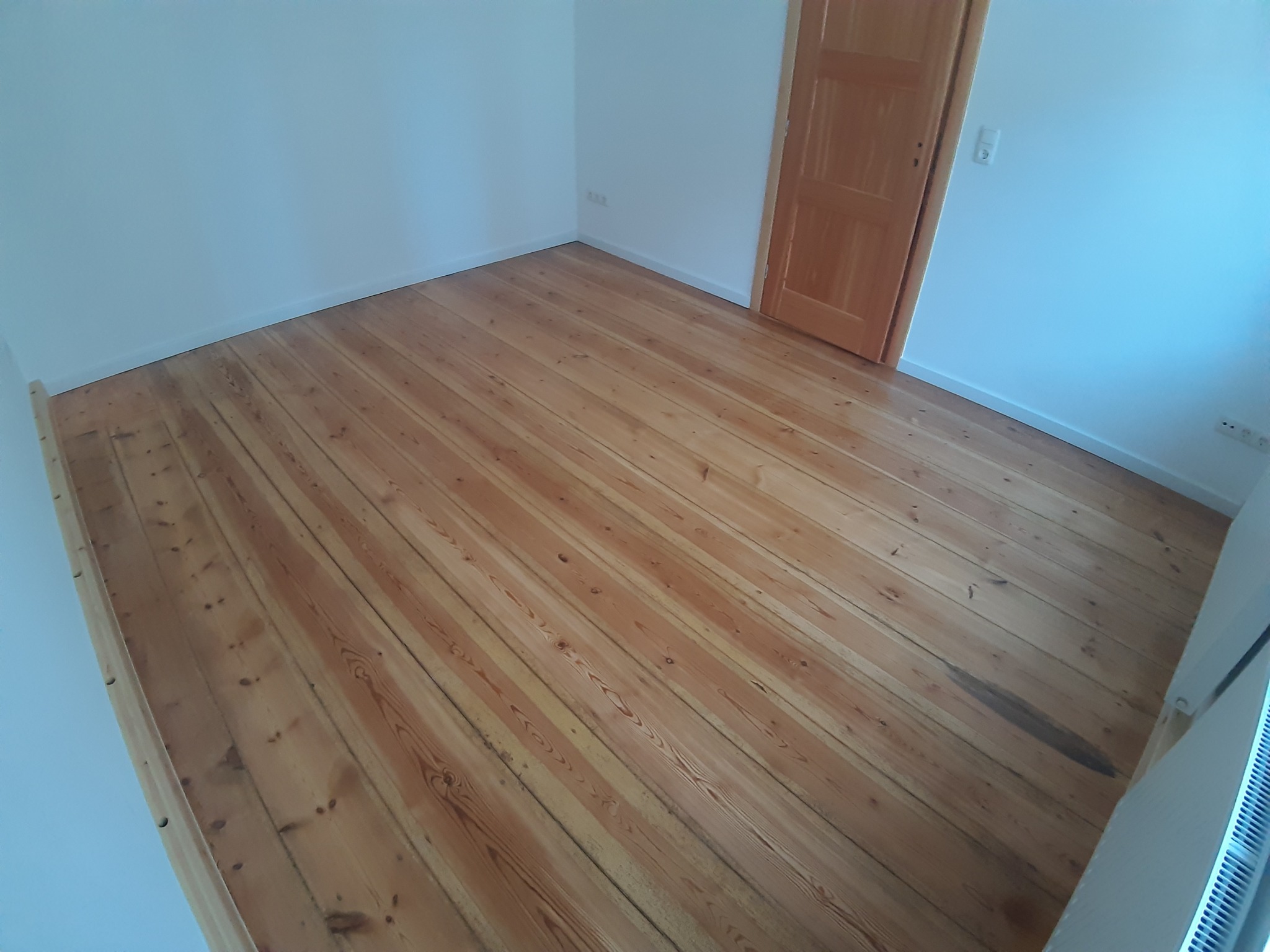 Jasne, drewniane deski podłogowe ułożone w pokoju z białymi ścianami i drewnianymi drzwiami. Widoczne ślady użytkowania i naturalna struktura drewna.