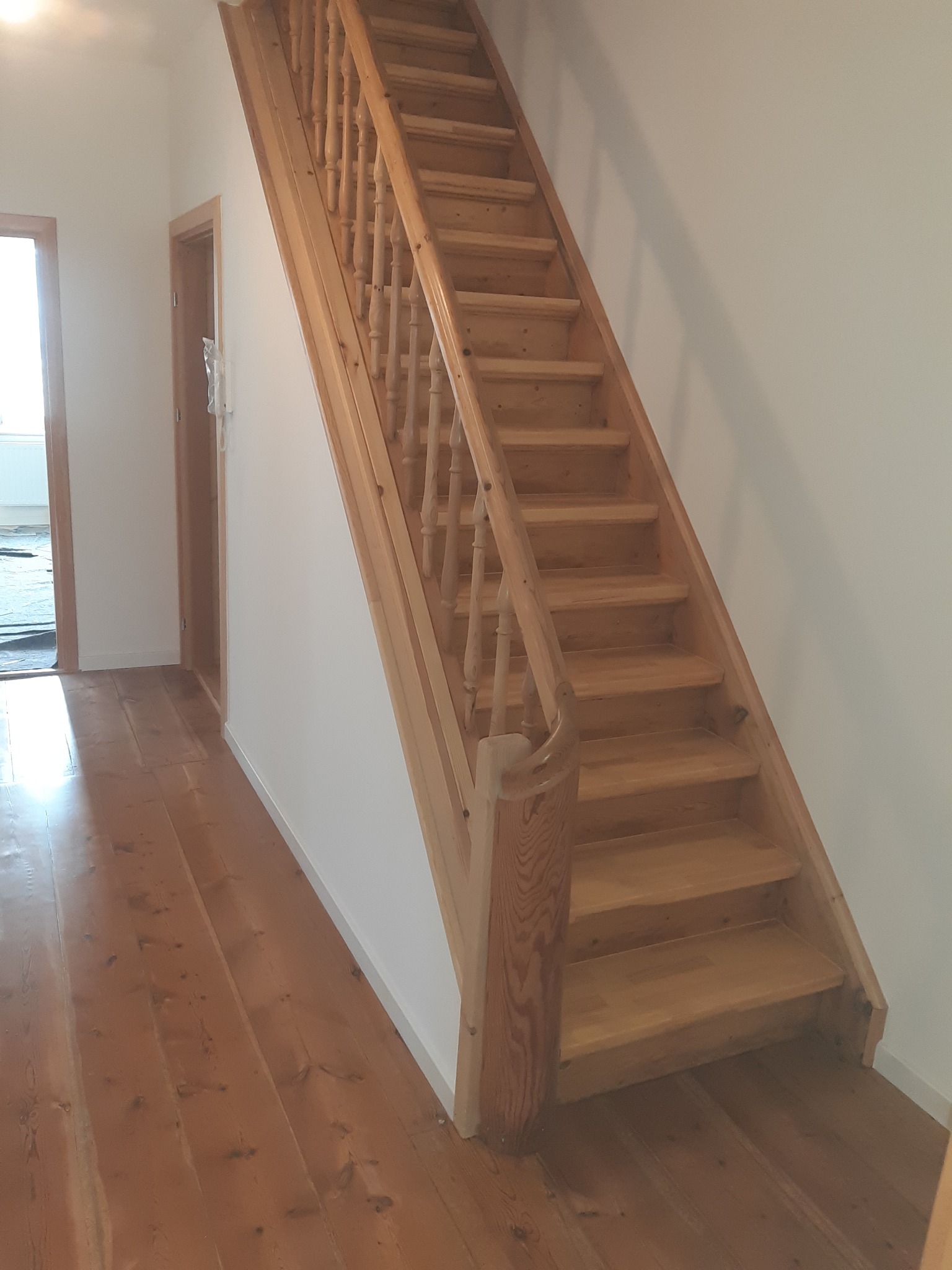 Jasne, drewniane schody z balustradą w korytarzu z drewnianą podłogą, widoczne drzwi po lewej stronie.