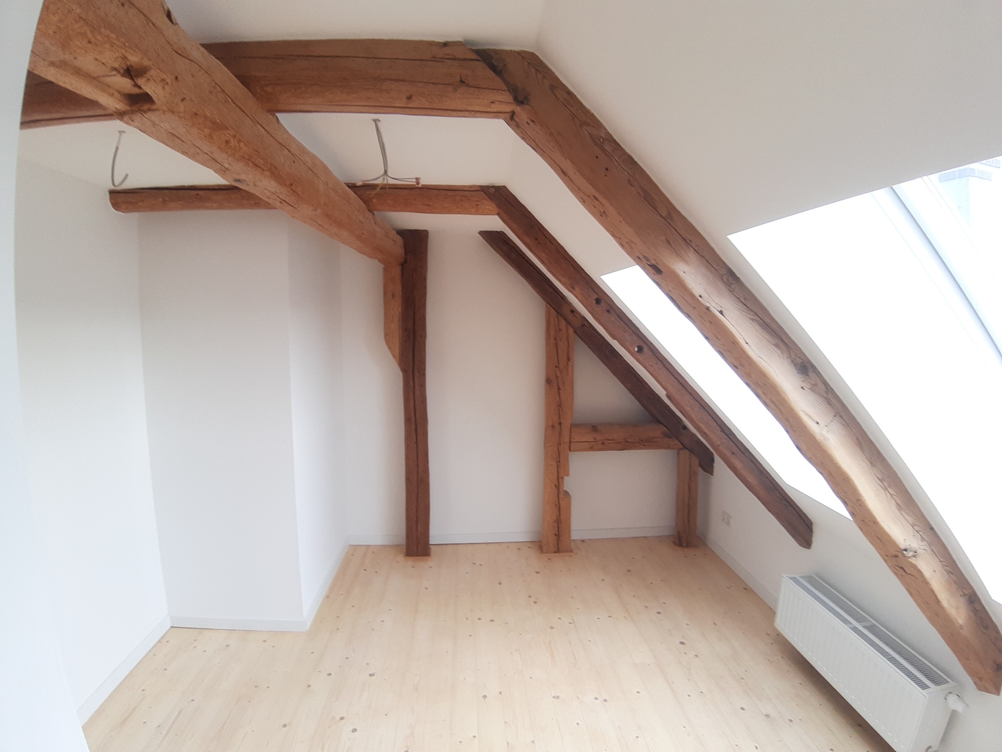 Poddasze z widocznymi elementami konstrukcyjnymi dachu: drewnianymi belkami nośnymi, słupami i jasną drewnianą podłogą. Ściany białe, z oknem dachowym wpadającym w kadr. Widoczny grzejnik pod oknem.