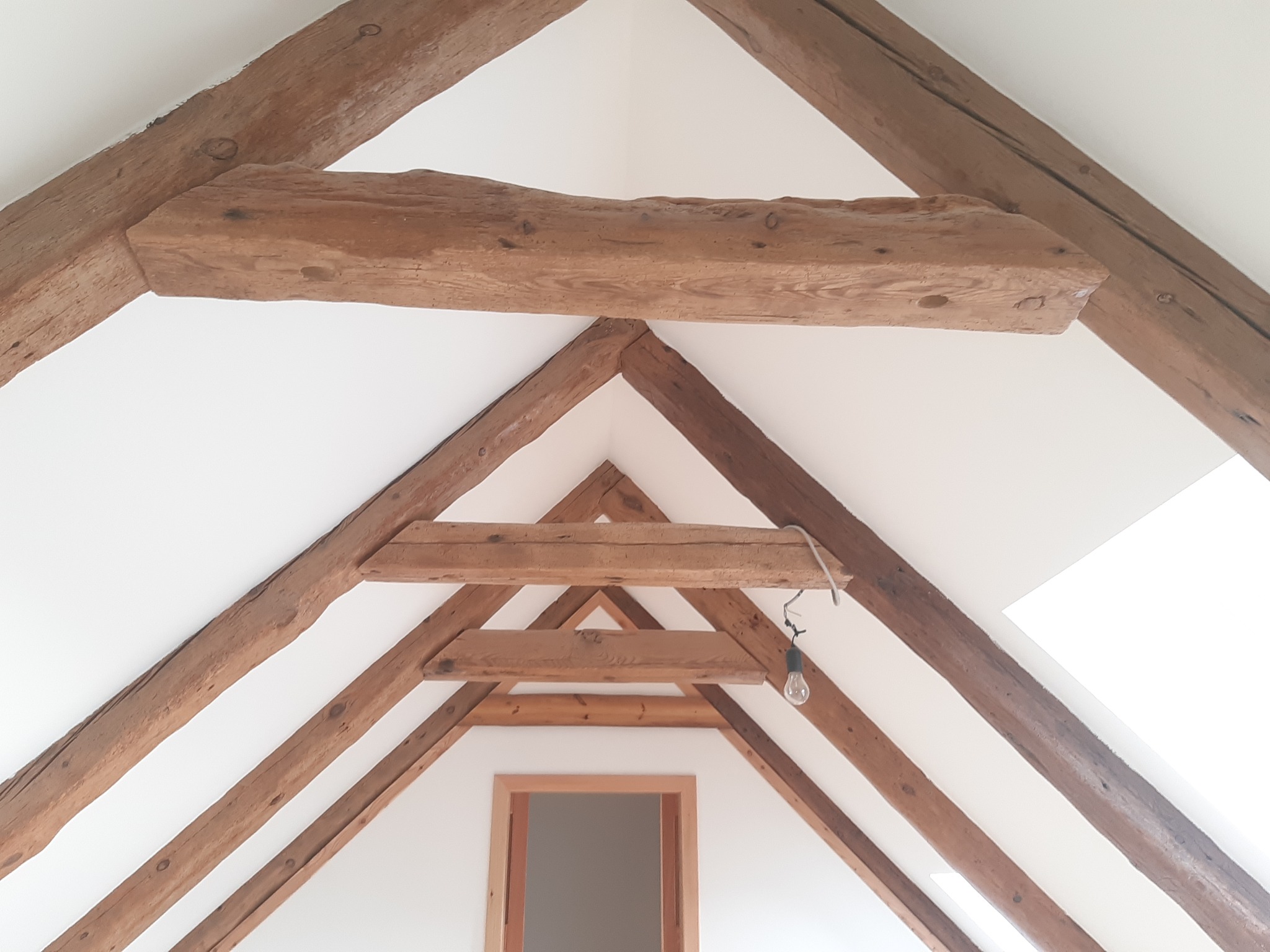 Drewniana konstrukcja dachu z widocznymi belkami w jasnym, odnowionym wnętrzu poddasza, z surowym wykończeniem i żarówką na kablu.