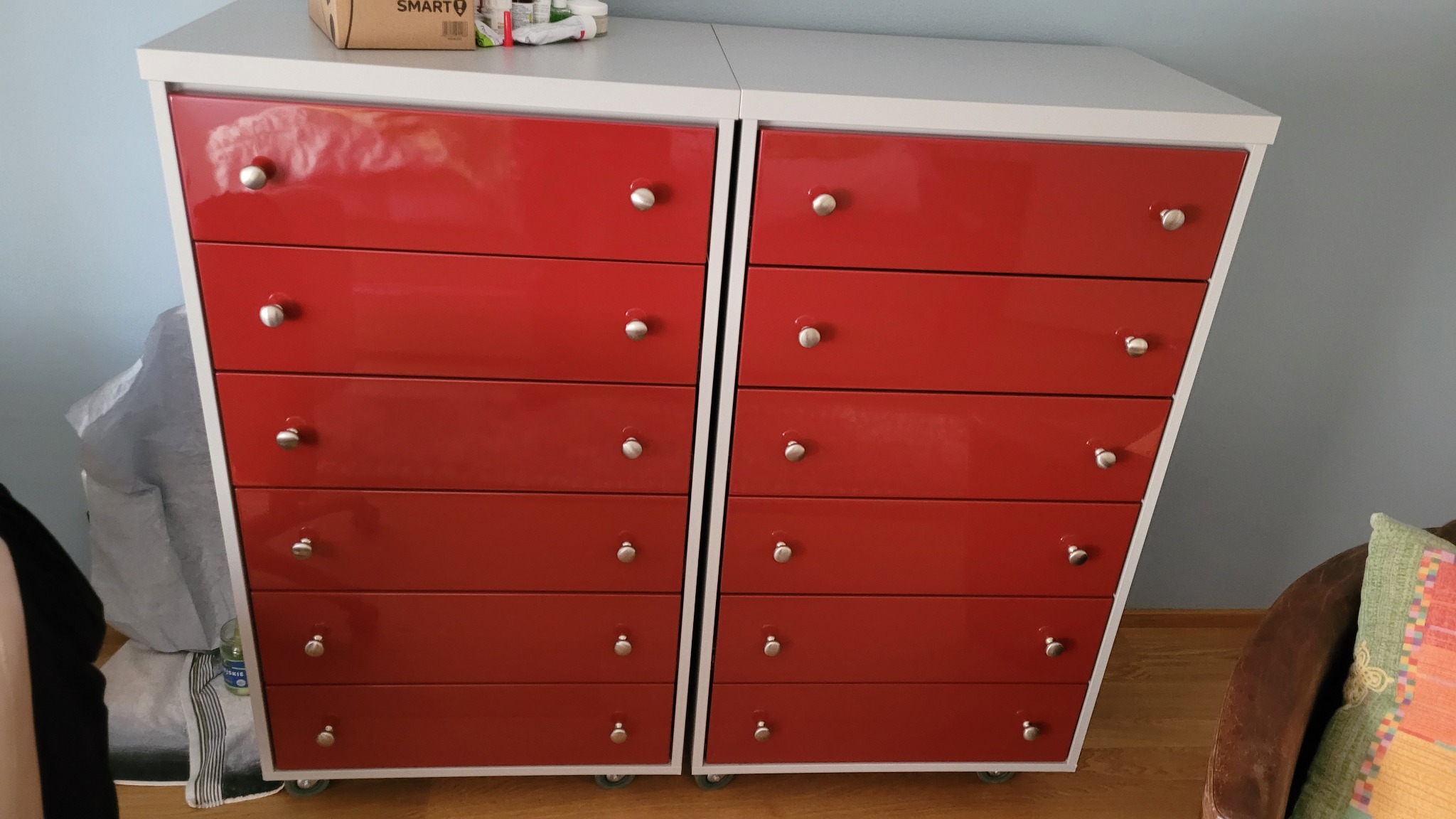 Dwie czerwone komody z sześcioma szufladami każda, z białymi blatami i bokami, na kółkach, z okrągłymi, srebrnymi uchwytami, ustawione obok siebie na drewnianej podłodze.