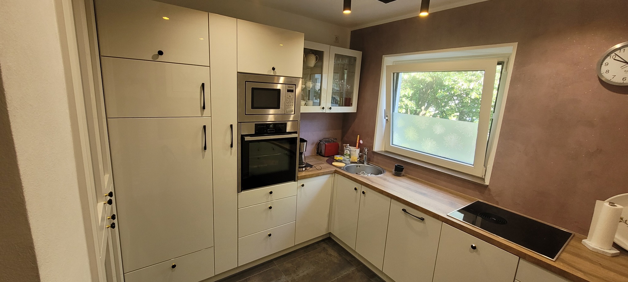 Jasna kuchnia z zabudową, zintegrowanym piekarnikiem i mikrofalówką, drewnianym blatem i oknem z dekoracyjną folią.