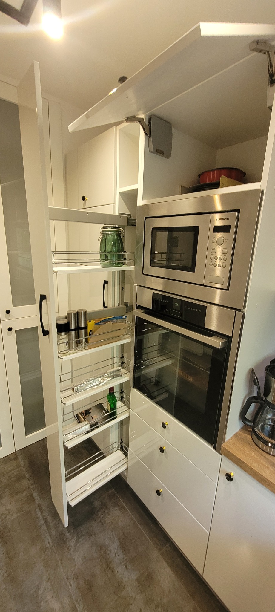 Biała kuchnia z wysuwaną szafką cargo, wyposażona w metalowe półki z przyprawami, słoikiem i butelką, z piekarnikiem i mikrofalówką w zabudowie.