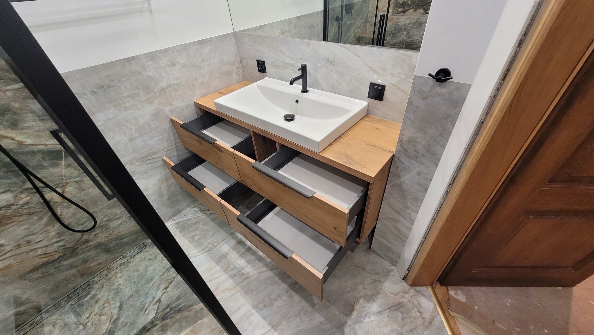 Nowoczesna szafka łazienkowa z drewnianym blatem i umywalką nablatową; otwarte szuflady z szarymi wnętrzami; marmurowe płytki na ścianach i podłodze; widoczny fragment kabiny prysznicowej...