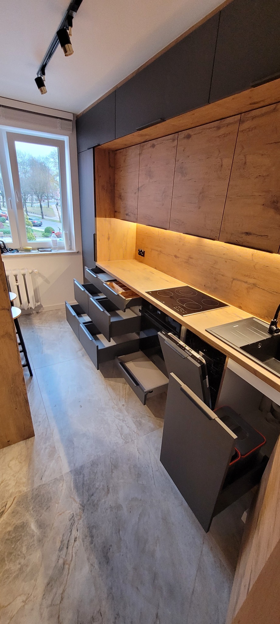 Nowoczesna kuchnia z drewnianymi frontami i szarymi szafkami, otwarte szuflady z organizerami, widoczna płyta indukcyjna i zlew, oświetlenie LED podszafkowe, marmurowa podłoga.