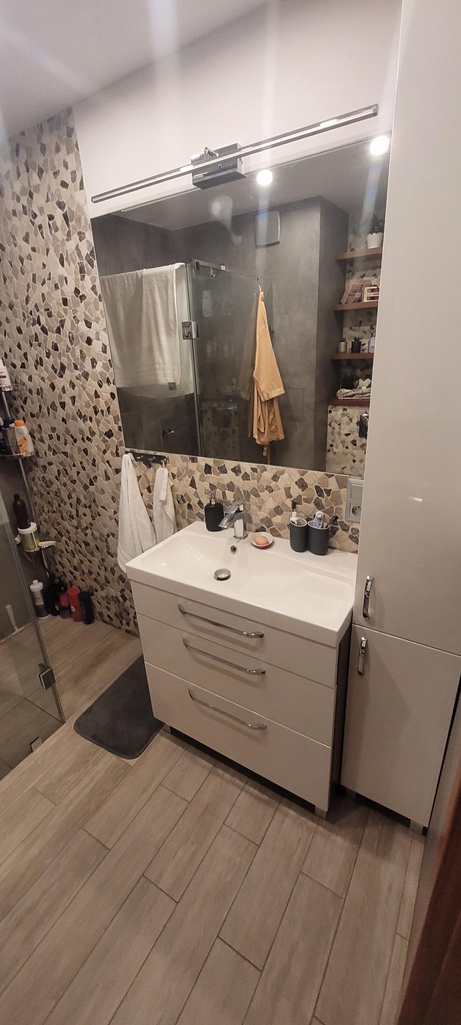 Nowoczesna łazienka z umywalką z szafką z trzema szufladami, wysoką szafką boczną, lustrem z oświetleniem i mozaikową ścianą w odcieniach beżu i brązu.