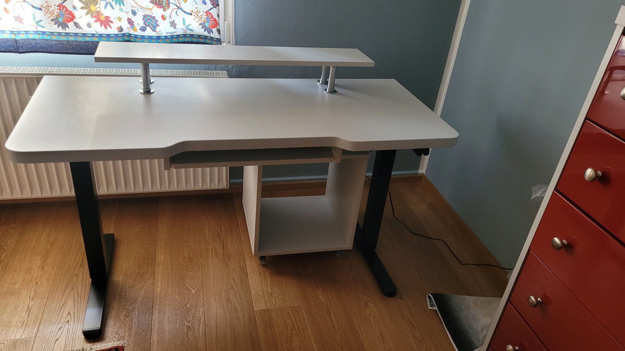 Białe biurko komputerowe z czarnymi nogami i wysuwaną półką na klawiaturę, dodatkową półką nad blatem, ustawione na drewnianej podłodze, obok czerwonej komody z szufladami.