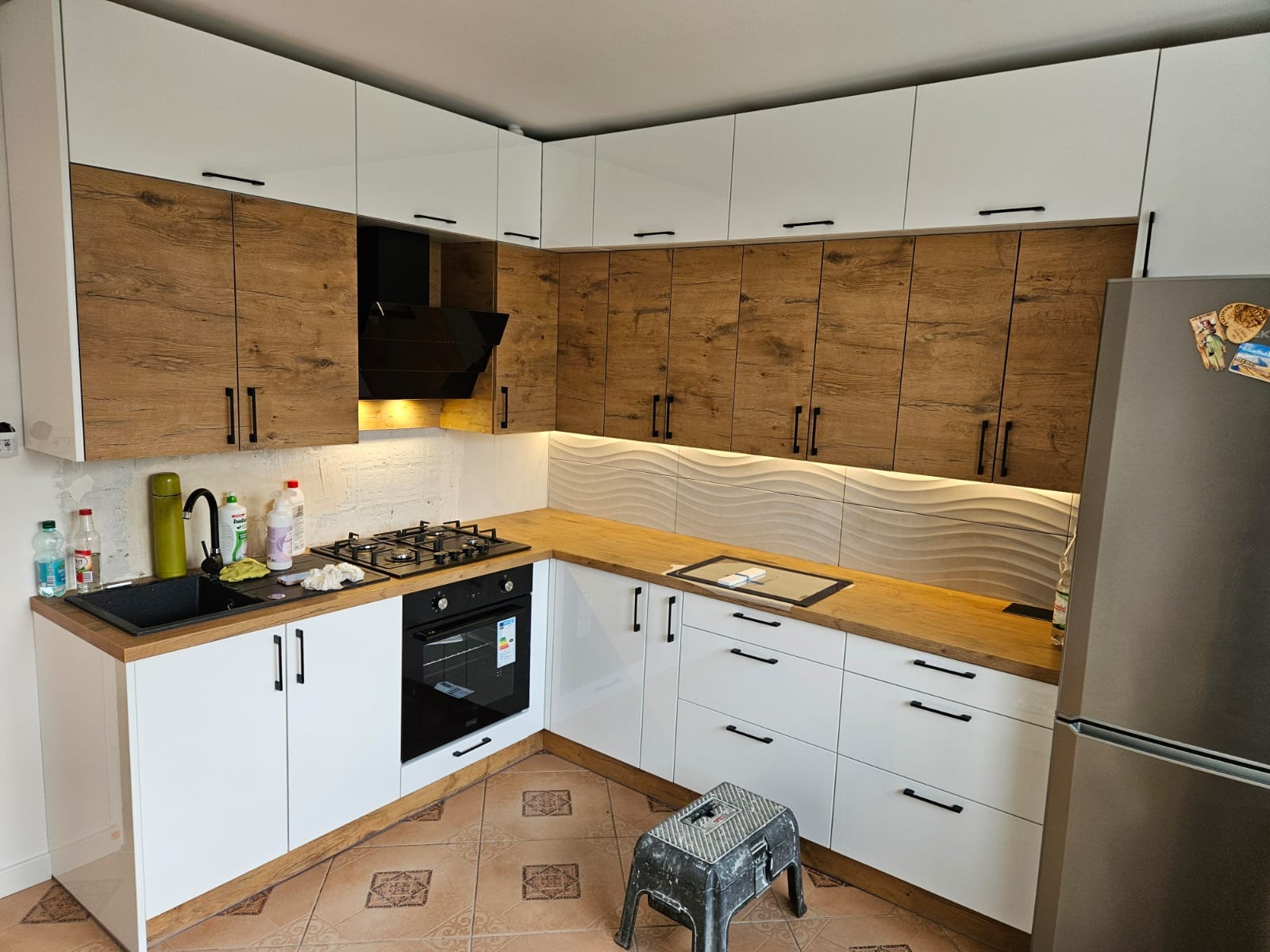 Nowoczesna kuchnia narożna z białymi szafkami dolnymi i górnymi szafkami w kolorze drewna, czarne uchwyty, drewniany blat, płyta gazowa i piekarnik. Ściana nad blatem wykończona dekoracyjnymi...