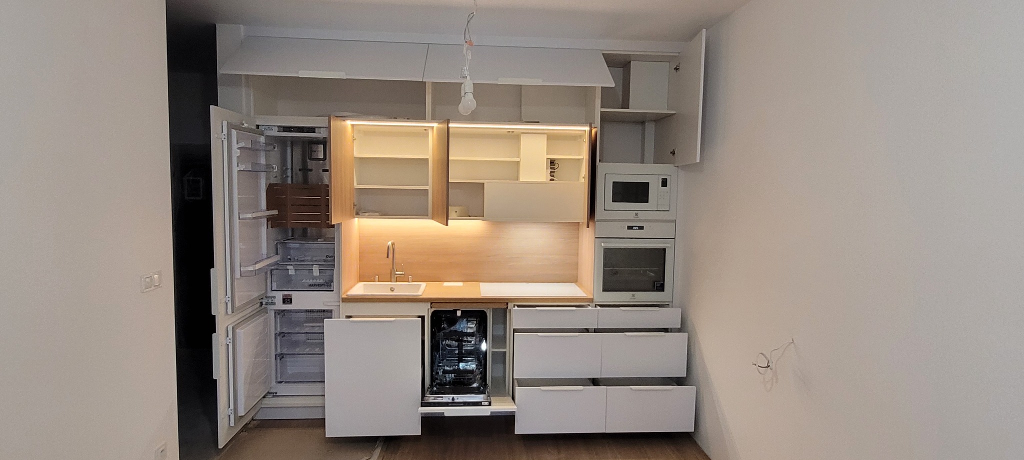 Biała zabudowa kuchenna z otwartą lodówką, zlewem, i zmywarką, oświetleniem LED pod szafkami wiszącymi i sprzętami AGD wkomponowanymi w zabudowę, w trakcie montażu, widoczne wiszące kable.