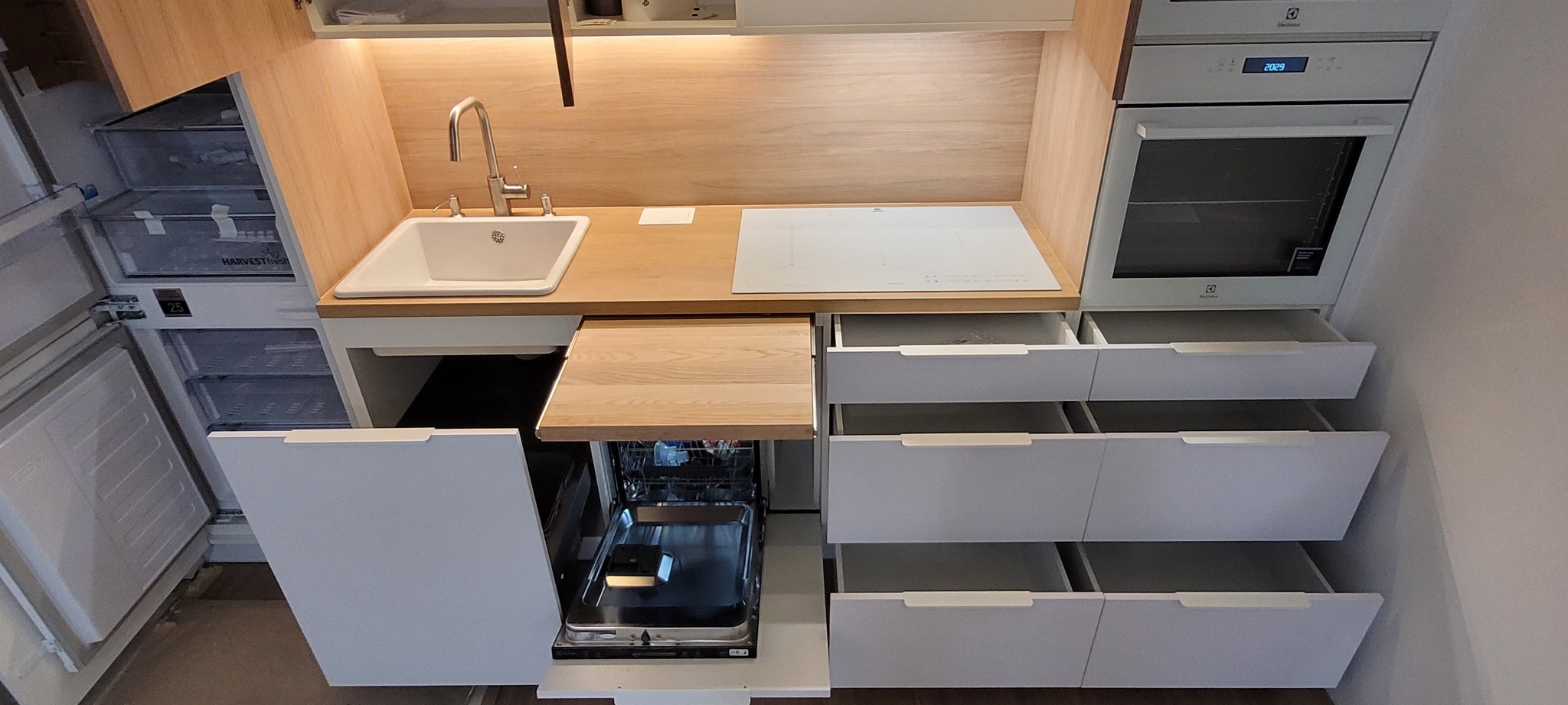 Nowoczesna, funkcjonalna zabudowa kuchenna z białymi frontami, drewnianym blatem, wbudowanym piekarnikiem Electrolux, płytą indukcyjną i otwartą zmywarką.