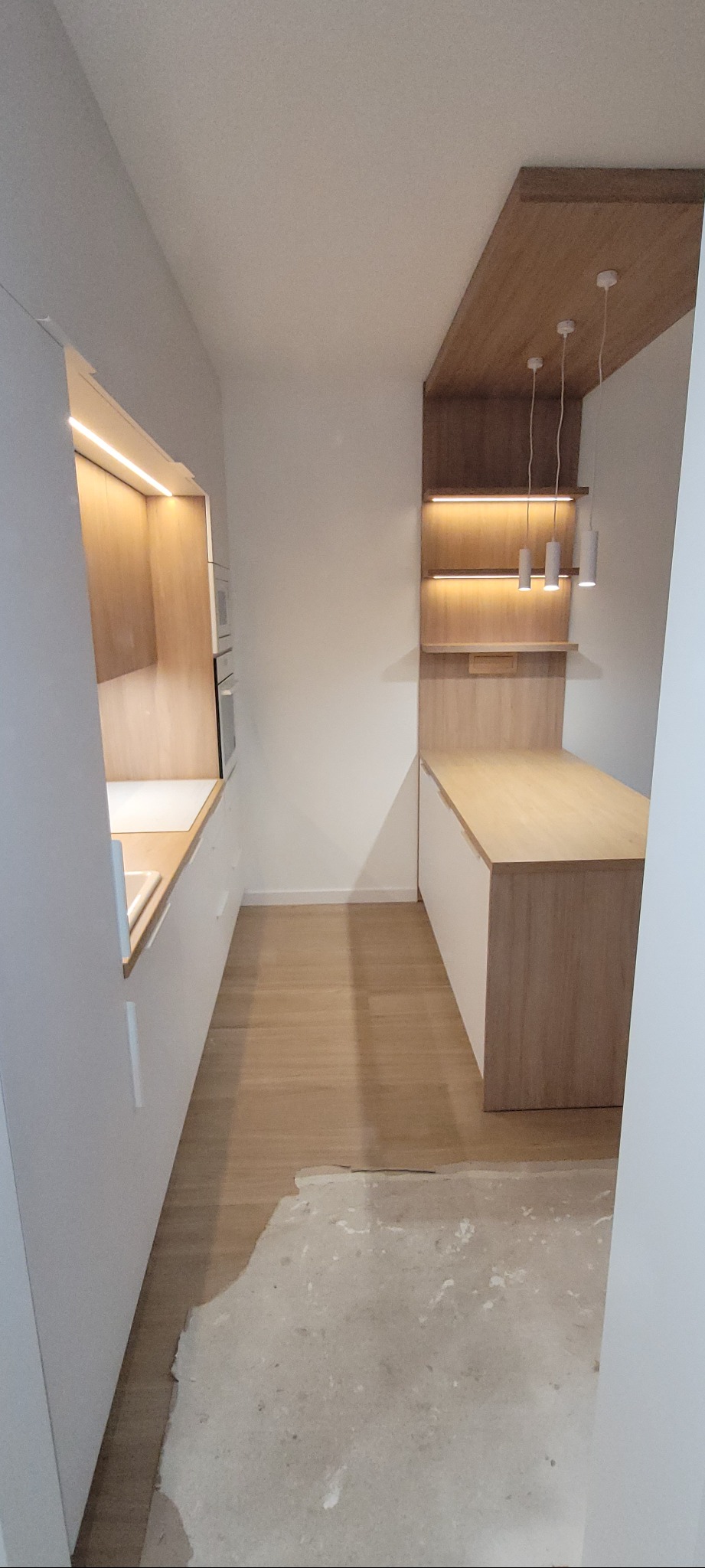 Wąska kuchnia z białymi szafkami i drewnianymi blatami, oświetleniem LED pod szafkami wiszącymi i trzema wiszącymi lampami cylindrycznymi, z niedokończoną podłogą.