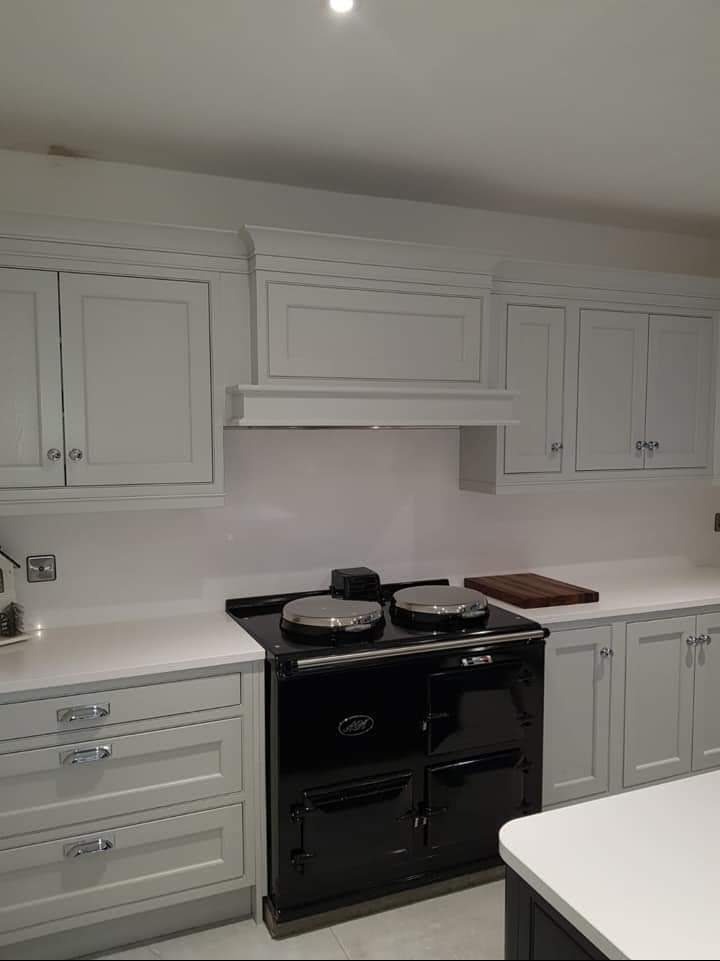 Jasnoszara kuchnia z czarnym piecem wolnostojącym, srebrne pokrętła i uchwyty, biały blat, oświetlenie punktowe.